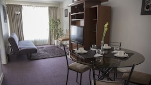 Un coin repas avec une table ronde, des chaises et un téléviseur. On aperçoit un canapé et une plante en pot à l’arrière-plan.