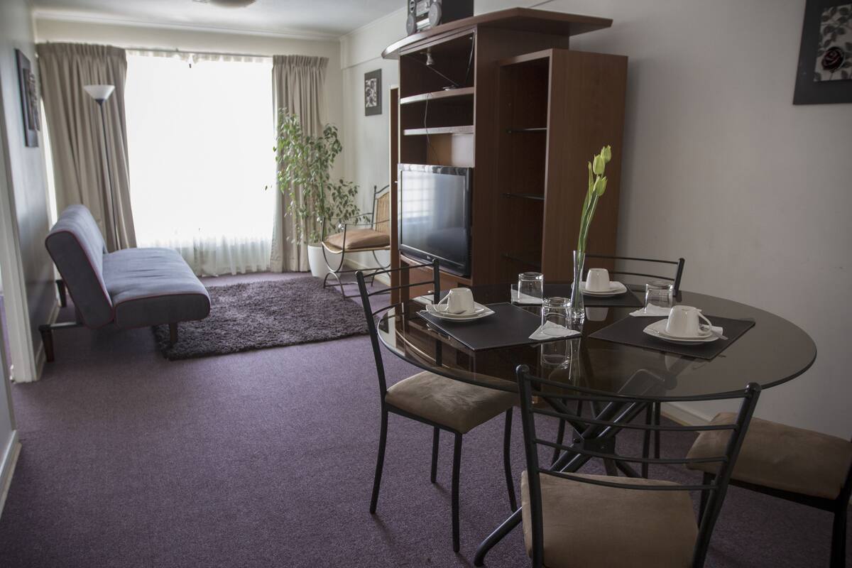 A dining area with a round table, chairs, and a television. There is a sofa and a potted plant in the background.