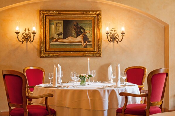 A dining area with a table set for four, red upholstered chairs, a framed painting above the table, and wall sconces with candles.
