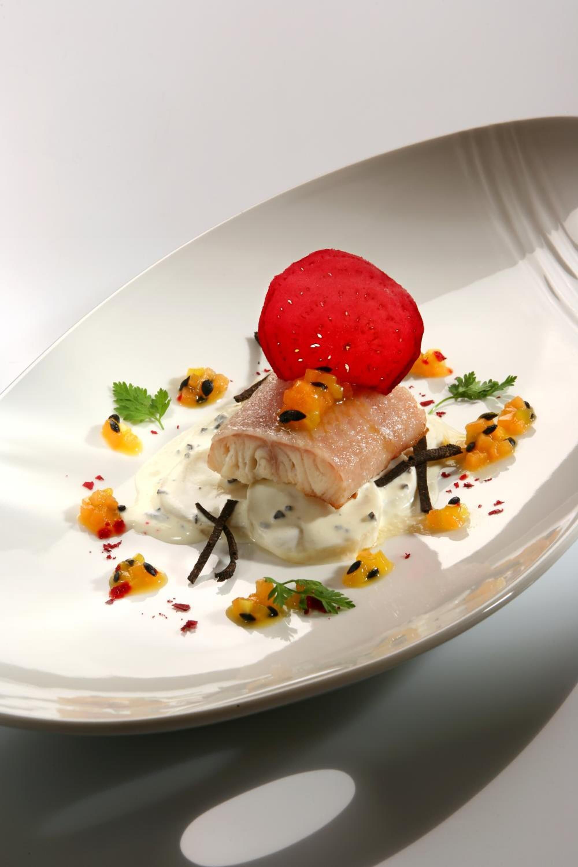 A white plate with a piece of fish, mango, and a red fruit on a white background.