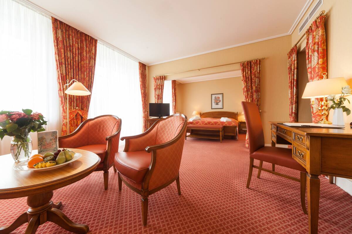 Een hotelkamer met een bed, twee fauteuils, een bureau, een tafel met fruit en gordijnen.