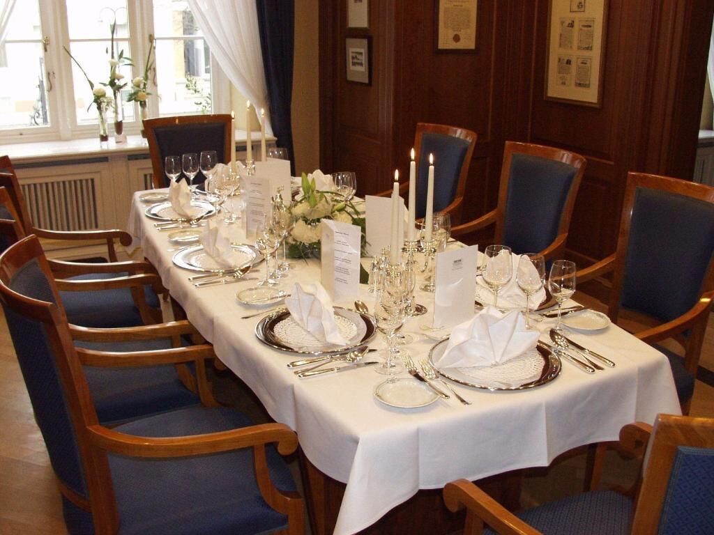 Mesa formal para comedor con mantel blanco, cubiertos y velas, rodeada de sillas tapizadas en azul en una habitación con paneles de madera.