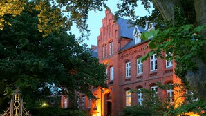 A historic red brick building with arched windows and a prominent entrance, surrounded by lush greenery.