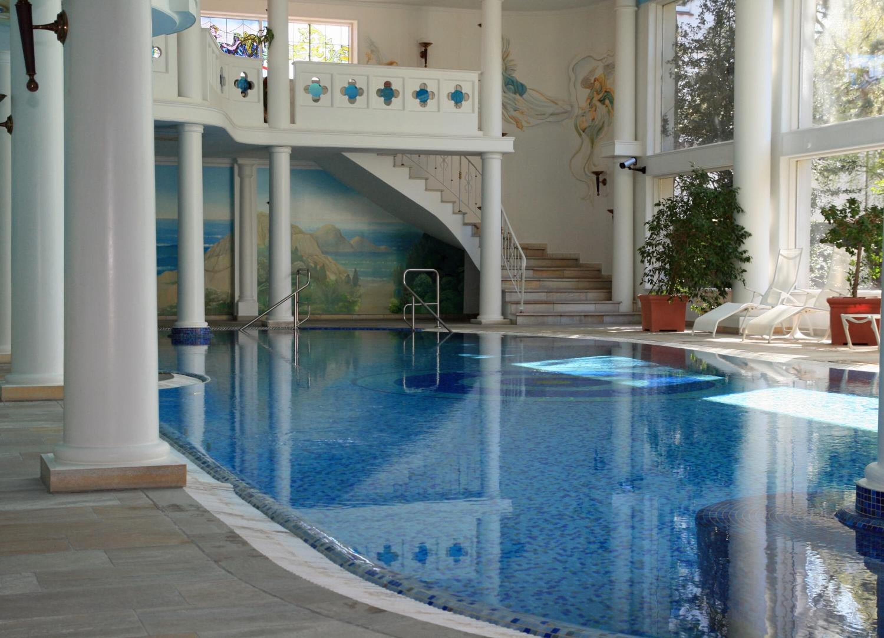 Une piscine intérieure à l’eau claire, un escalier menant à un balcon et des plantes en pot disposées autour de la piscine.