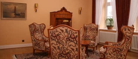 A room with four ornate chairs, a wooden table, a chandelier, a painting, and a window with curtains.
