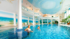 An indoor swimming pool with a mural of a beach scene, white columns, and lounge chairs.