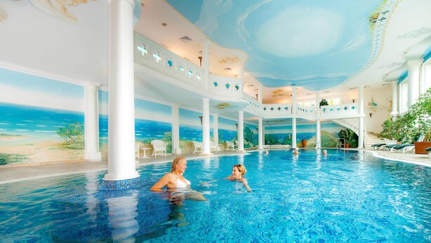 An indoor swimming pool with a mural of a beach scene, white columns, and lounge chairs.