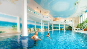 An indoor swimming pool with a mural of a beach scene, white columns, and lounge chairs.