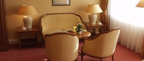 Habitación de hotel con sofá, dos sillones, una mesita, dos lámparas, un florero y un cuadro en la pared.