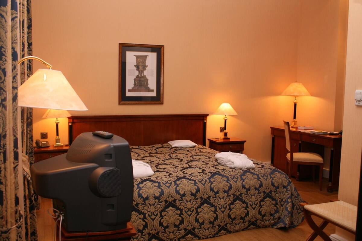 Hotelkamer met een bed, twee bedlampen, een bureau met stoel, een televisie en een lijst aan de muur.