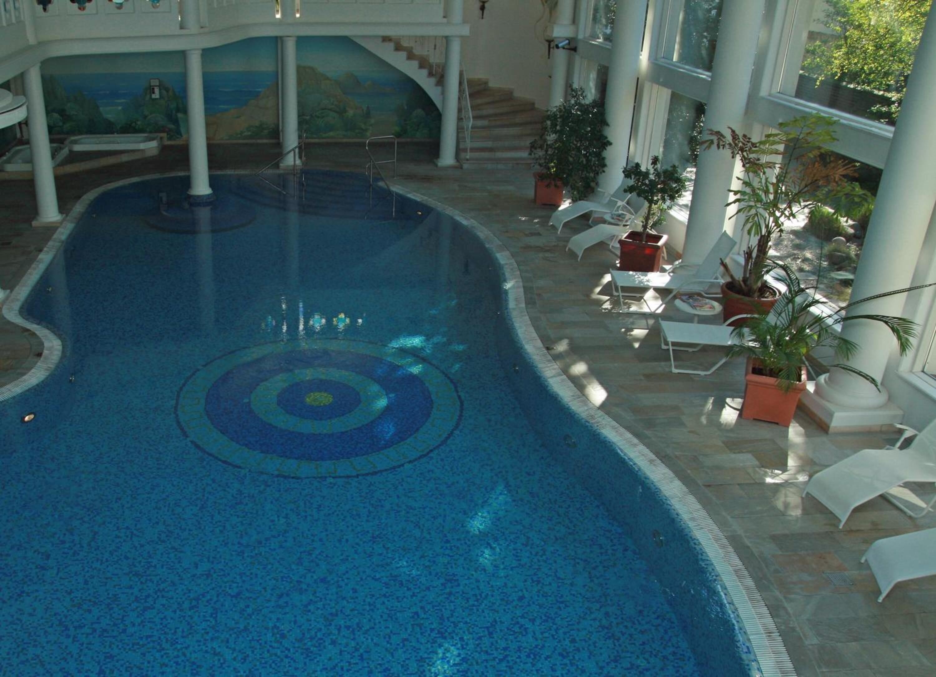 An indoor swimming pool with a central circular design, surrounded by lounge chairs and potted plants.
