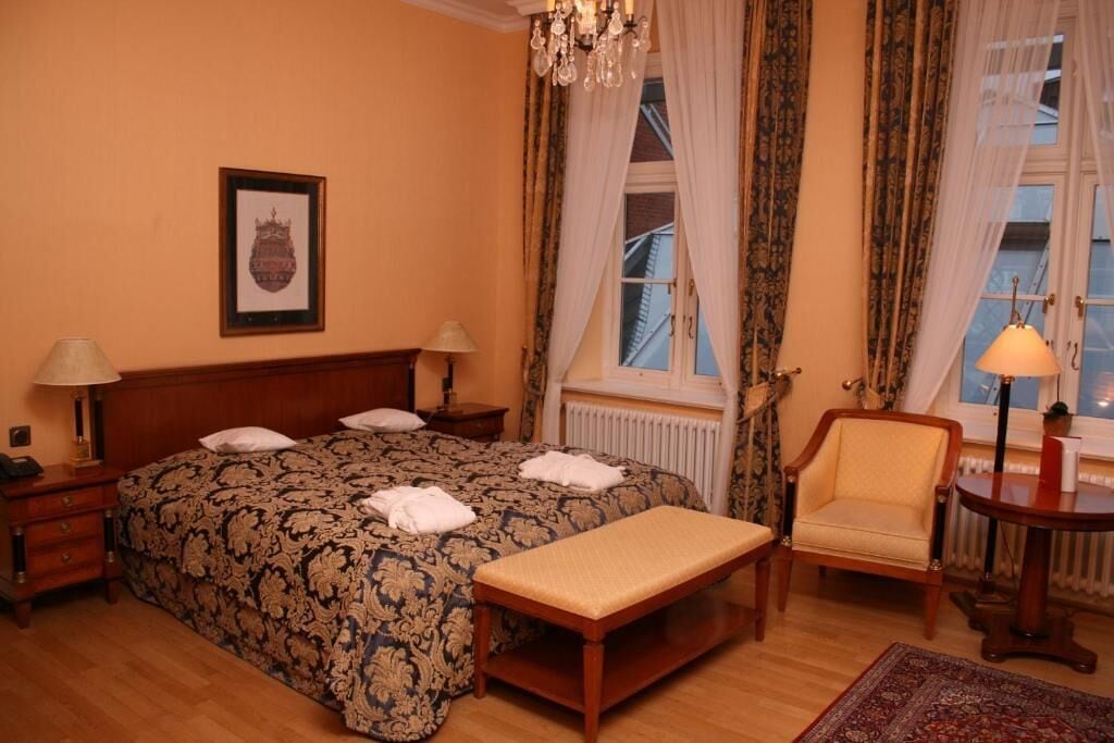 Une chambre d’hôtel avec un grand lit, deux tables de chevet, une chaise, un banc et une petite table avec une lampe.