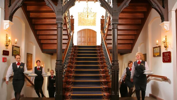 A grand staircase with ornate railings and a chandelier, leading to a high-ceilinged room.