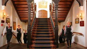 A grand staircase with ornate railings and a chandelier, leading to a high-ceilinged room.