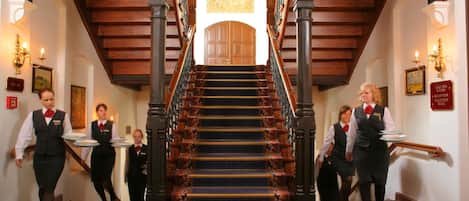 Una imponente escalera con barandillas ornamentales y un candelabro, que conduce a un salón con techos altos.