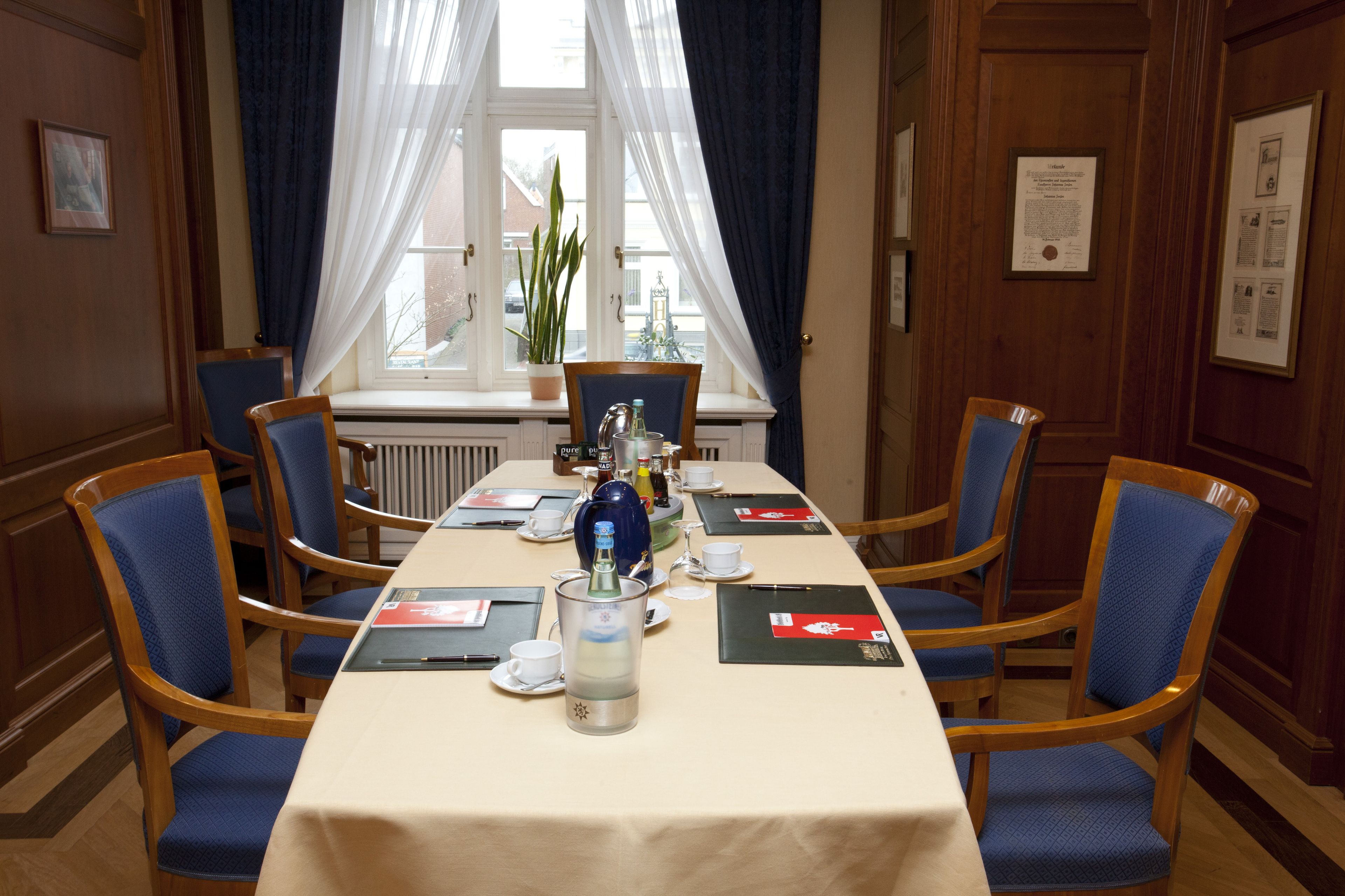 A conference room with a long table, blue chairs, a potted plant, and a window with curtains.