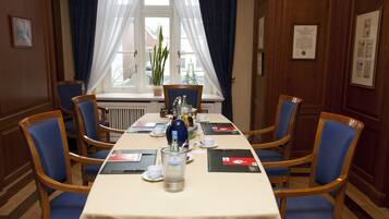 A conference room with a long table, blue chairs, a potted plant, and a window with curtains.