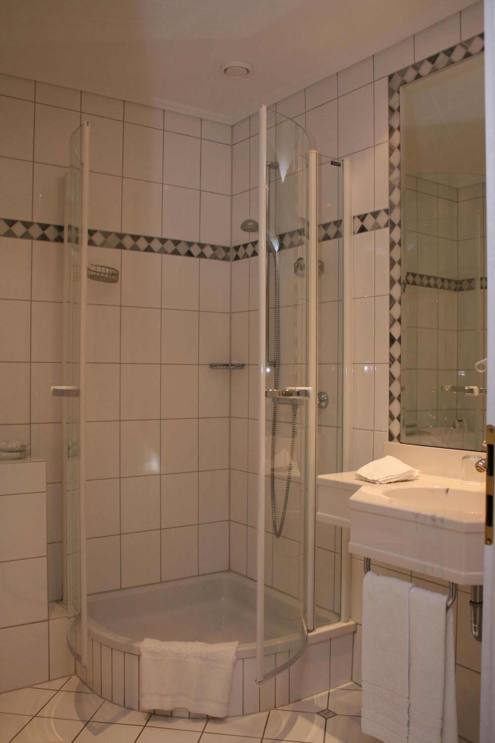A corner shower with a glass door, a white tiled wall, and a corner sink with a mirror.