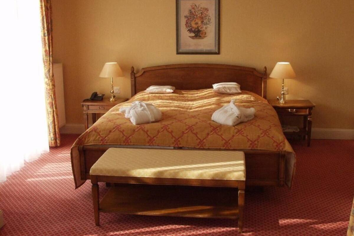 Een net opgemaakt bed met een houten hoofdbord, twee nachtkastjes met lampen en een lijst met bloemkunst op de muur.