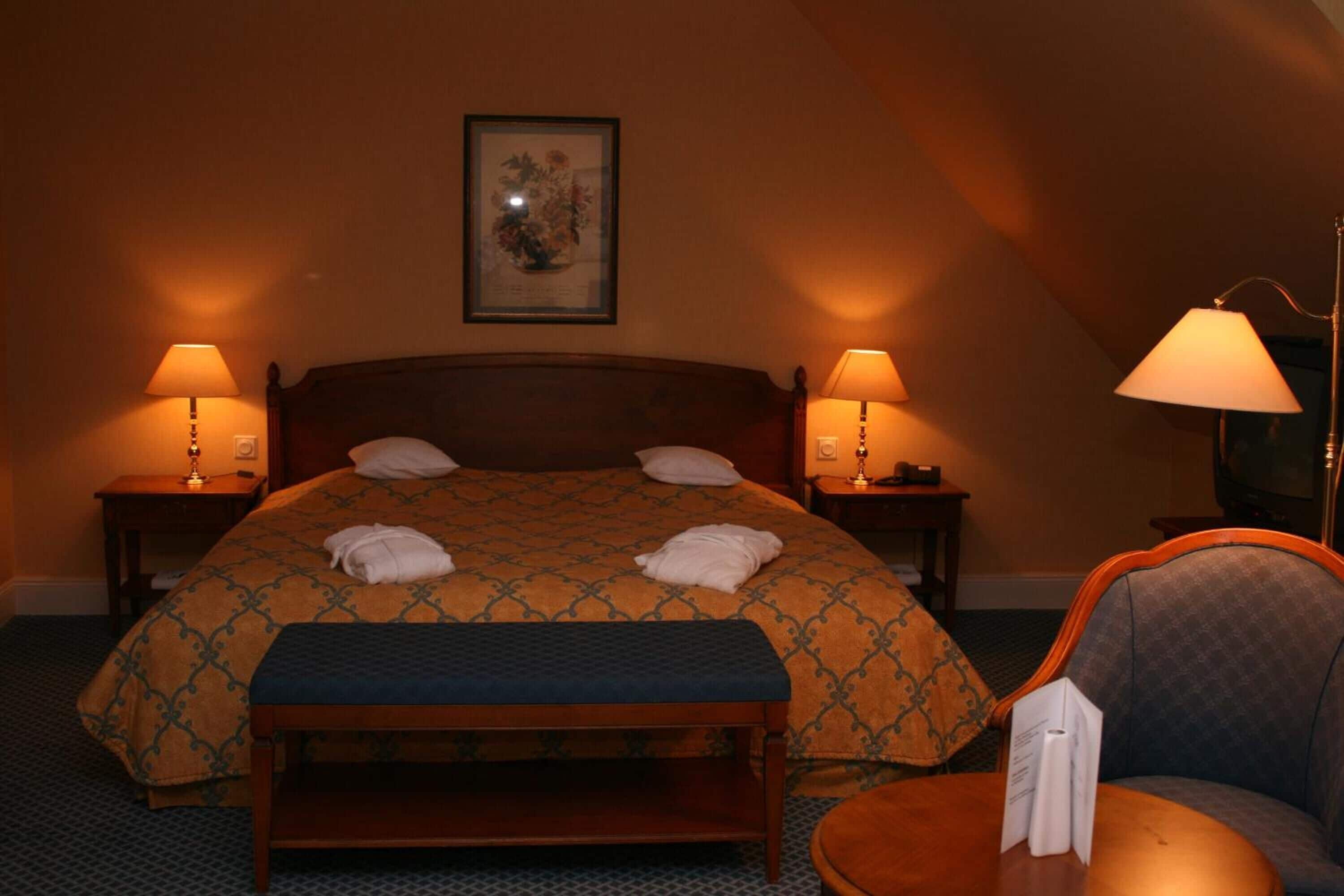 Een netjes opgemaakt bed met een bedsprei met patroon, twee bedlampen, een ingelijste foto aan de muur en een klein tafeltje met een lamp.