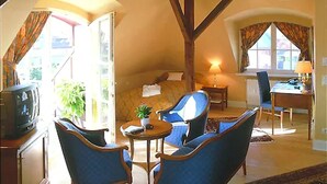 A cozy room with a bed, two blue armchairs, a small table, a desk with a chair, and a television.