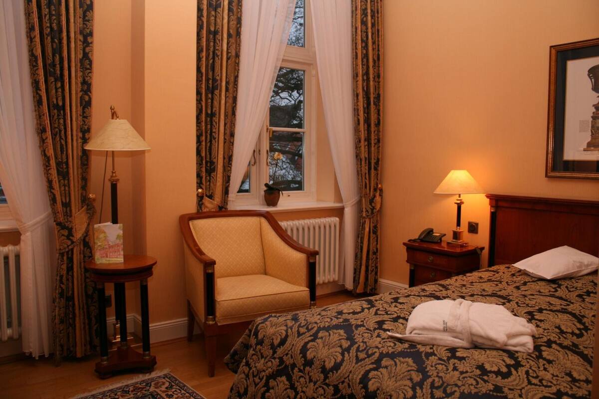 Een hotelkamer met een bed, een stoel, een lamp, een radiator en een raam met gordijnen.