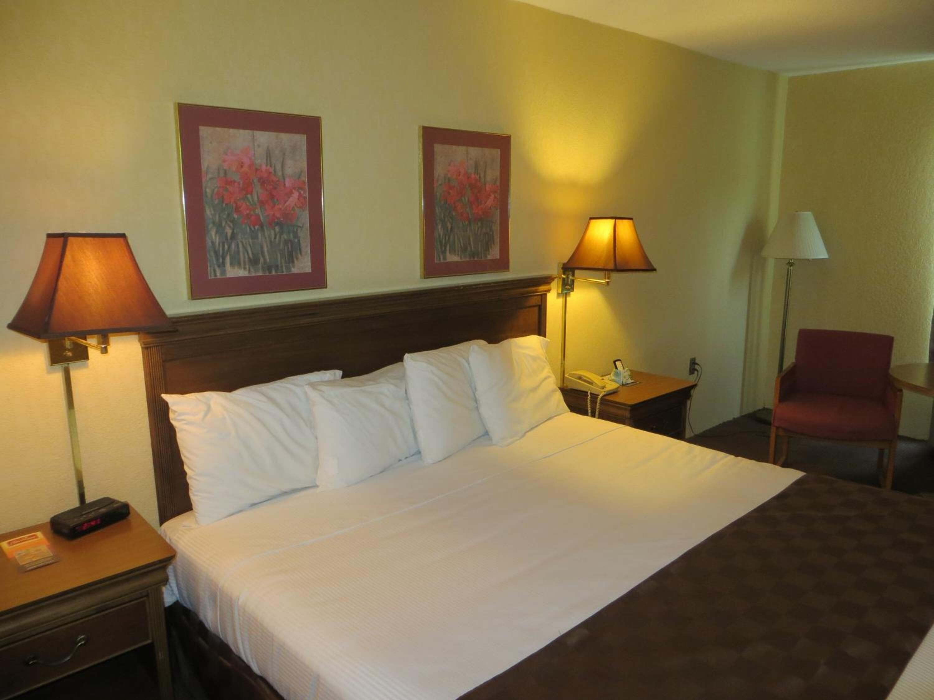 Una habitación de hotel con una cama grande, dos lámparas de noche, una silla y dos cuadros enmarcados con motivos florales en la pared.