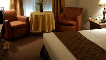 A hotel room with a large bed, two armchairs, a small table with a vase, and a lamp on a nightstand.