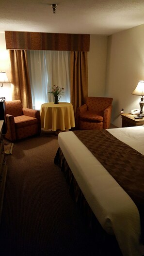 A hotel room with a large bed, two armchairs, a small table with a vase, and a lamp on a nightstand. - Americourt Hotel (Mountain City)
