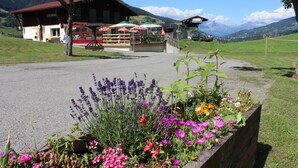 A building with a red roof, a wooden bench area, and a flower bed in the foreground. - Les Bernards (Praz-sur-Arly)