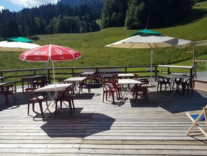 An outdoor patio with tables and chairs, surrounded by a wooden deck and green hills in the background. - Les Bernards (Praz-sur-Arly)