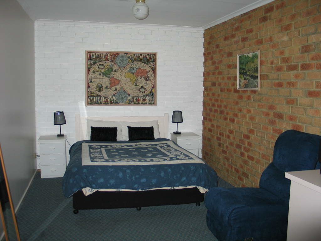 A bedroom with a bed, two bedside tables, a blue chair, a map on the wall, and a brick wall.