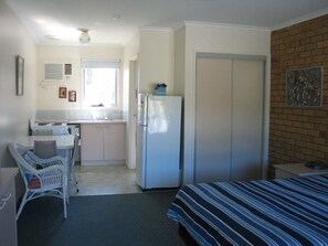 Une chambre d’hôtel compacte avec une kitchenette, un lit avec une literie à rayures, une chaise en osier et un mur en briques.