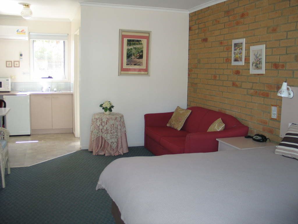 A hotel room with a bed, a red sofa, a small table with a floral tablecloth, and a kitchen area visible through an open door.