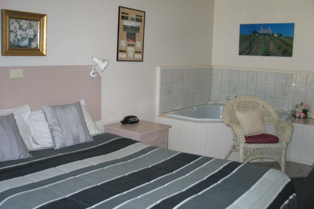 A bedroom with a bed, a nightstand, a chair, a wall-mounted lamp, and a picture of a landscape.
