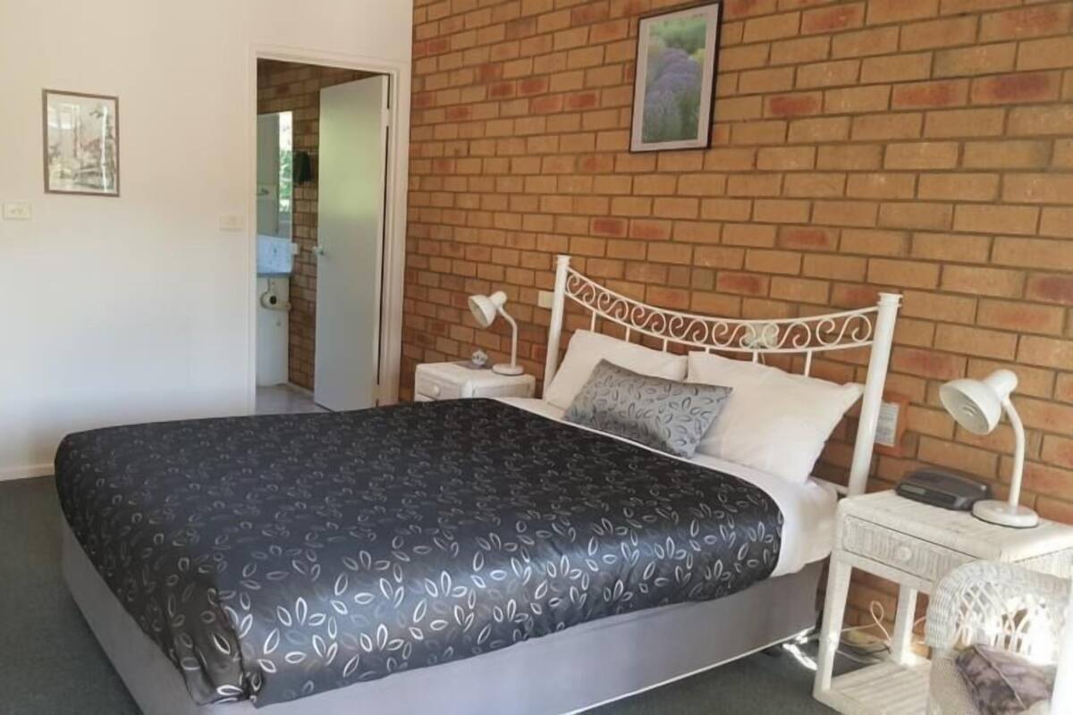 A bedroom with a brick wall, a bed with a patterned bedspread, two bedside tables with lamps, and a door leading to another room.