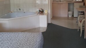 A room with a large bed, a bathtub, a small kitchen area, and a window with a view of greenery. - Balnarring Village Motor Inn (Balnarring)
