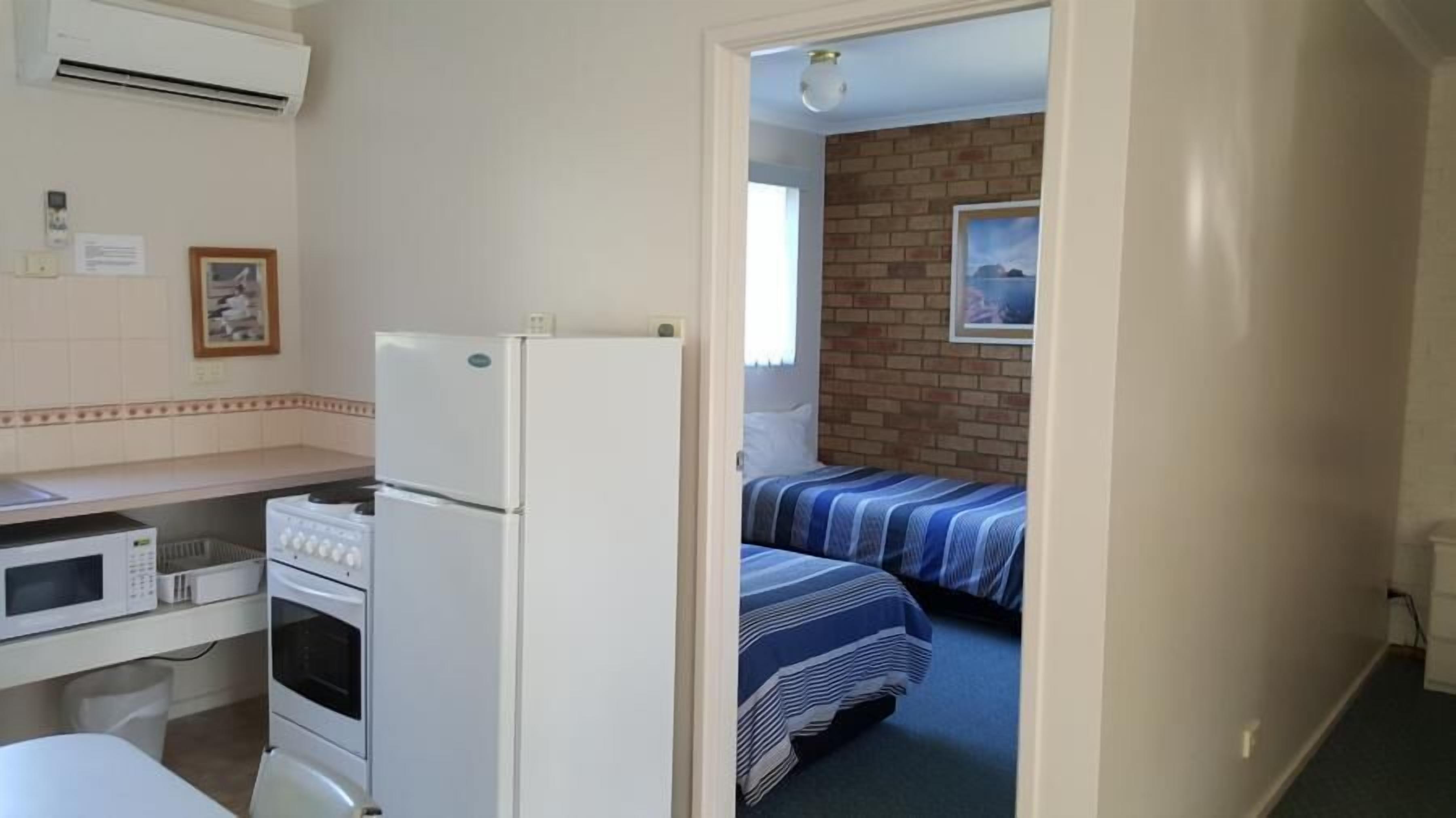 A compact kitchen with a refrigerator, stove, and microwave. A bed with striped bedding is visible in the adjacent room.