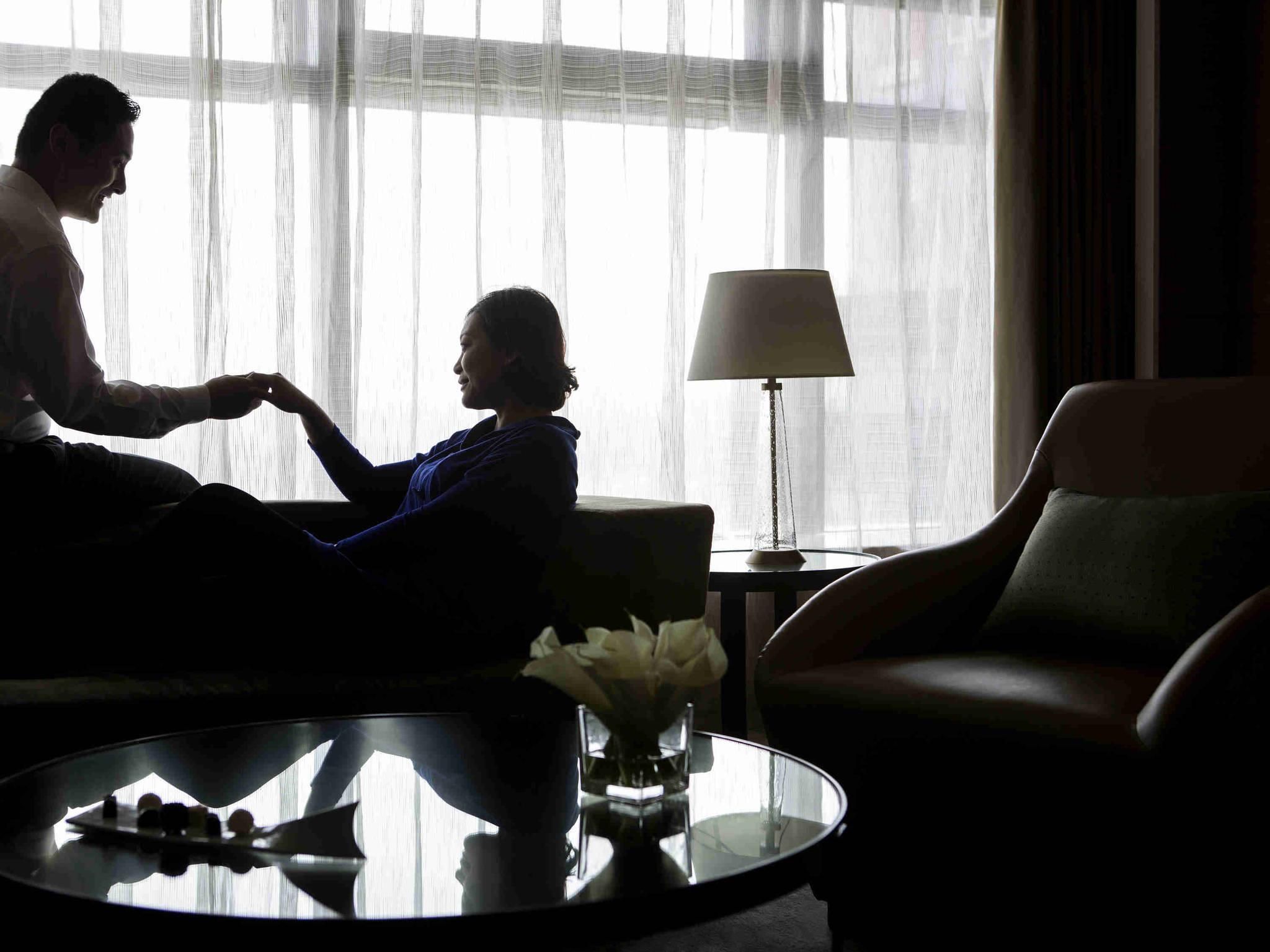 A dimly lit room with a person sitting on a couch, another person standing, and a table with a lamp and flowers.