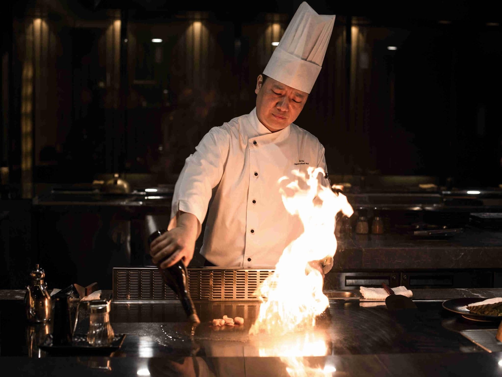 A chef in a white uniform with a tall hat, cooking on a flat-top grill with visible flames.