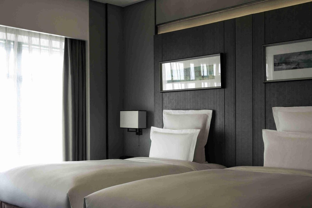 A hotel room with two beds, a bedside lamp, a framed picture on the wall, and a window with curtains.