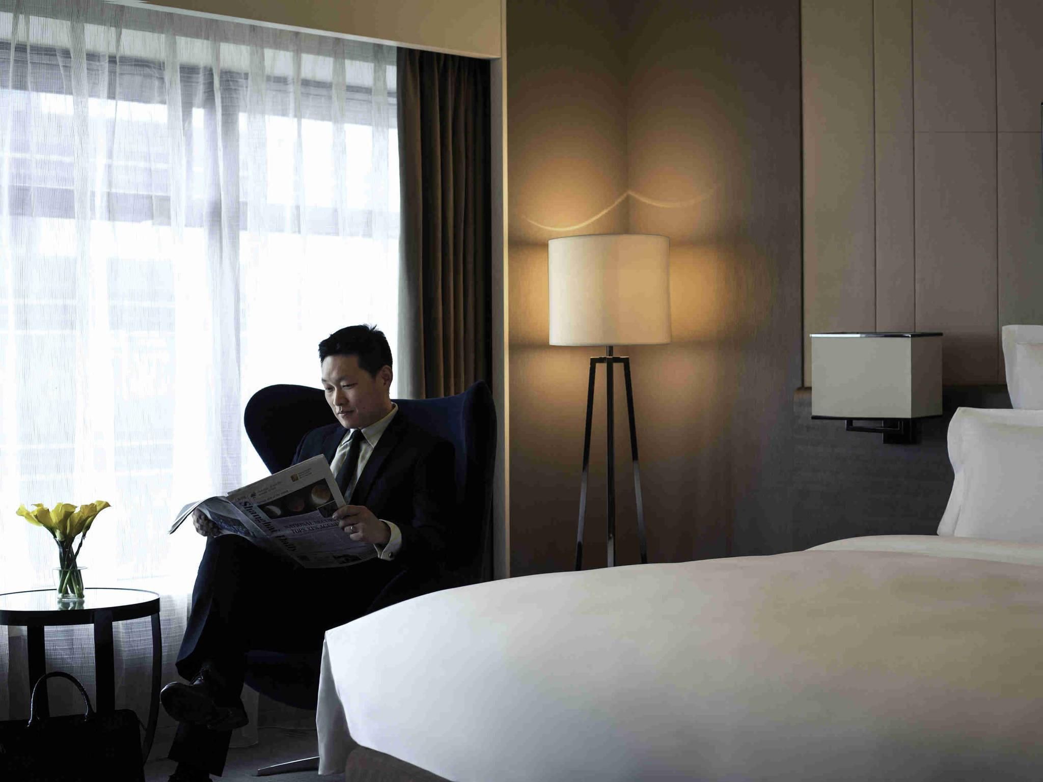 A hotel room with a bed, a reading chair, a standing lamp, a small table with a vase, and a window with curtains.
