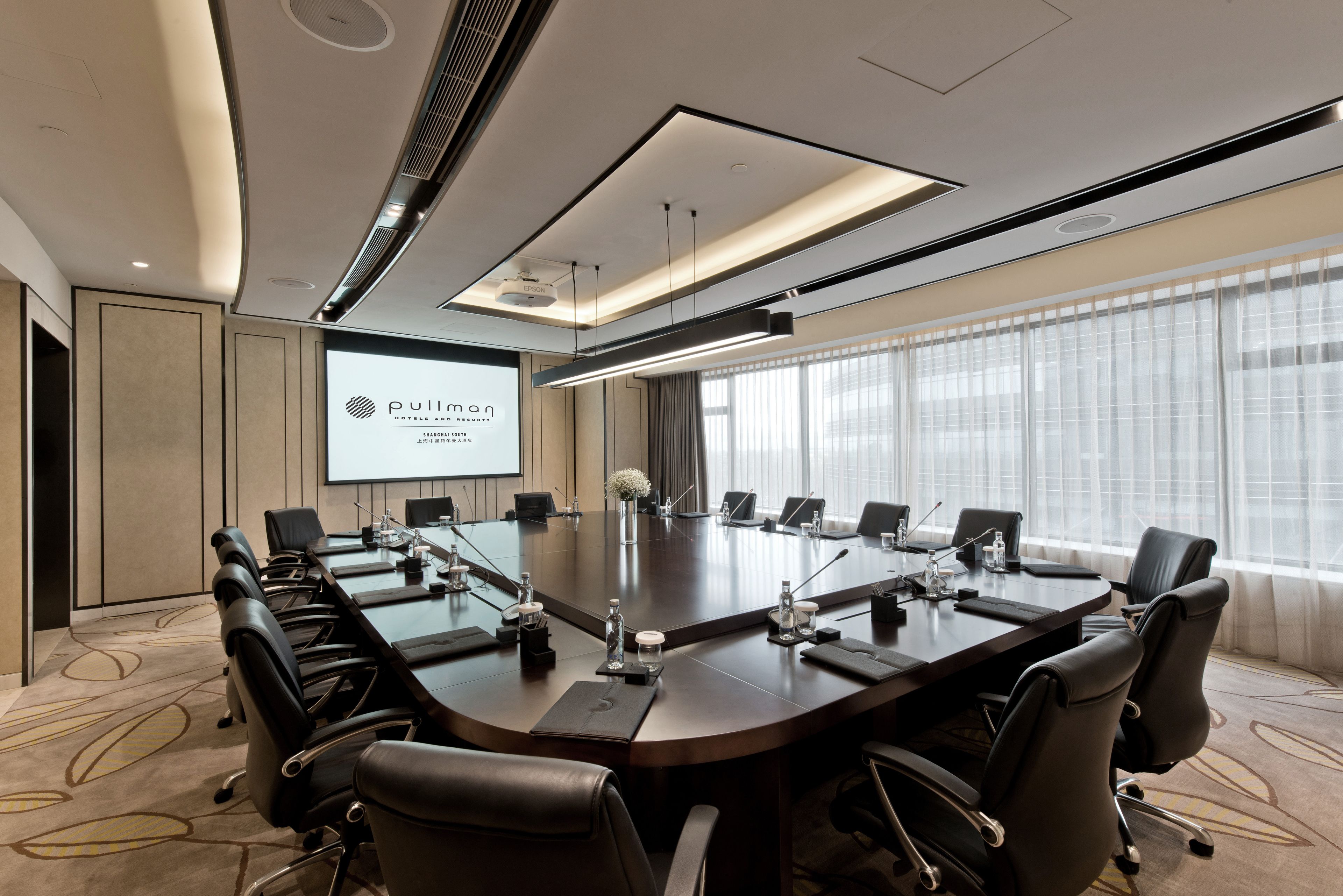 A conference room with a large table, chairs, a projector screen, and a large window with curtains.