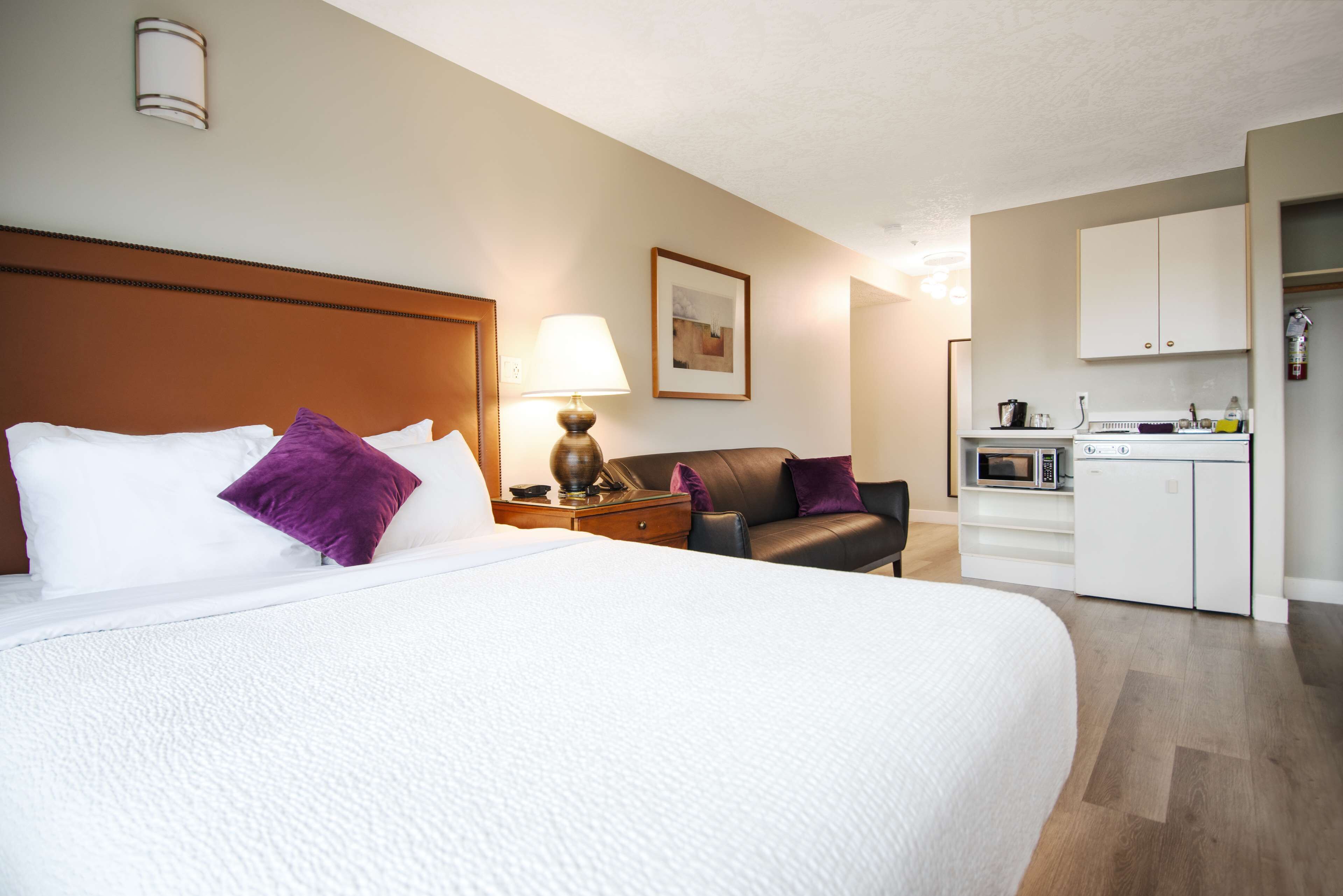 Habitación de hotel con una cama grande, un escritorio con lámpara, una silla, microondas y una pequeña kitchenette.
