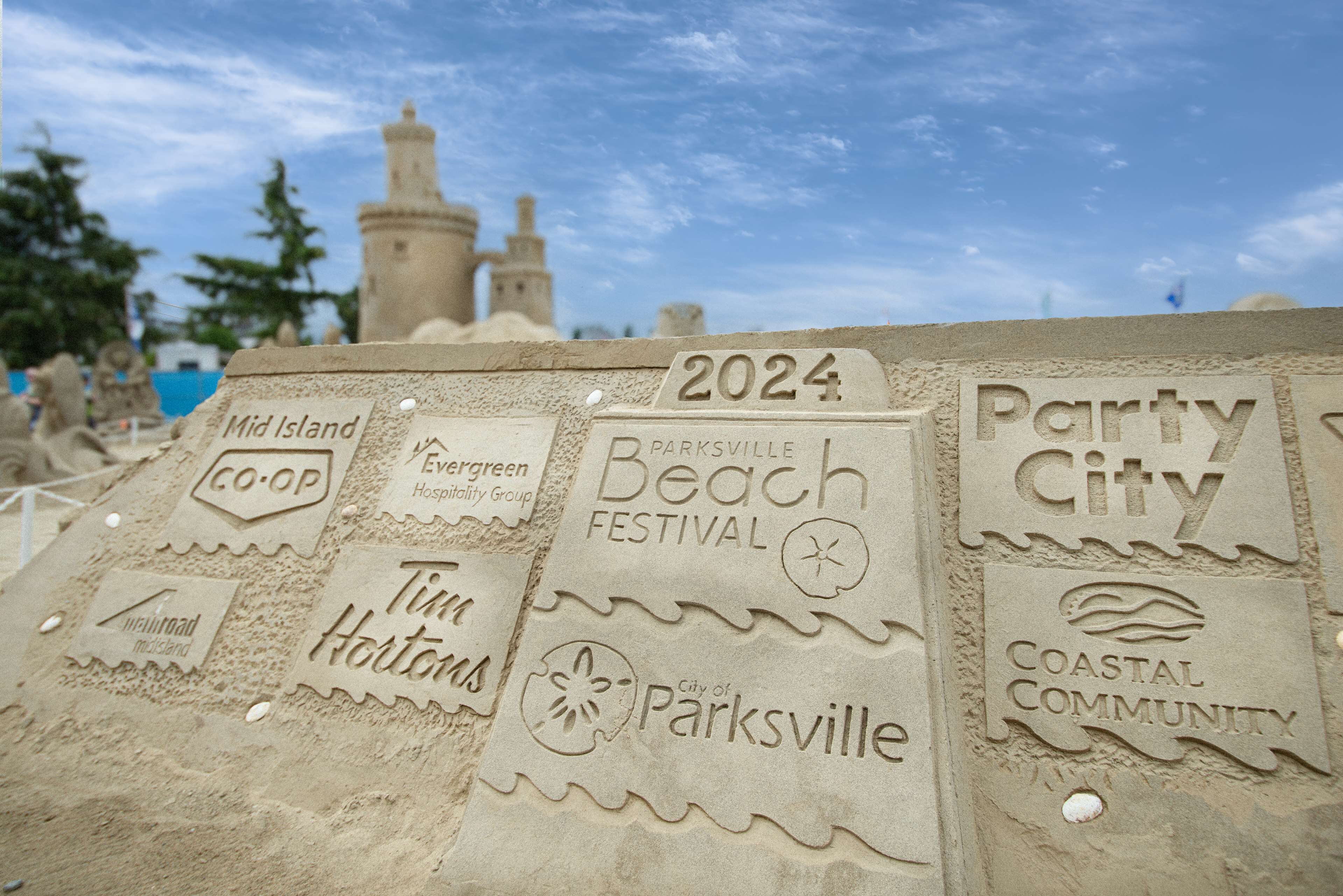 En sandskulptur med text som visar att det kommer att hållas en strandfestival år 2024, och som bland annat innehåller namnen Mid Island, Evergreen och Parksville.