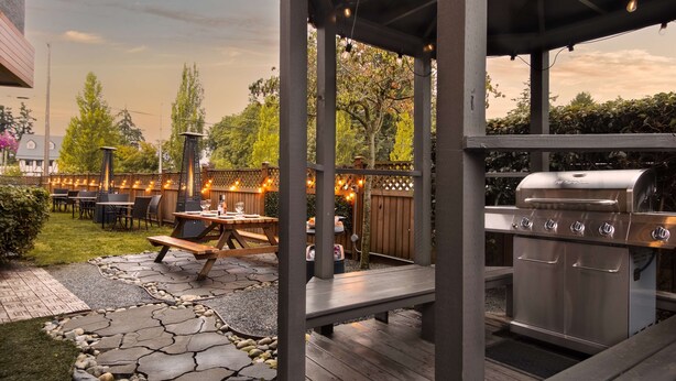 Eine Terrasse im Freien mit Steinplattenboden, Holztischen und einem fest eingebauten Grill.