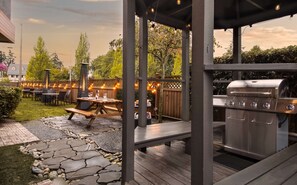 Een terras buiten met een vloer van stenen tegels, houten tafels en een ingebouwde barbecue.