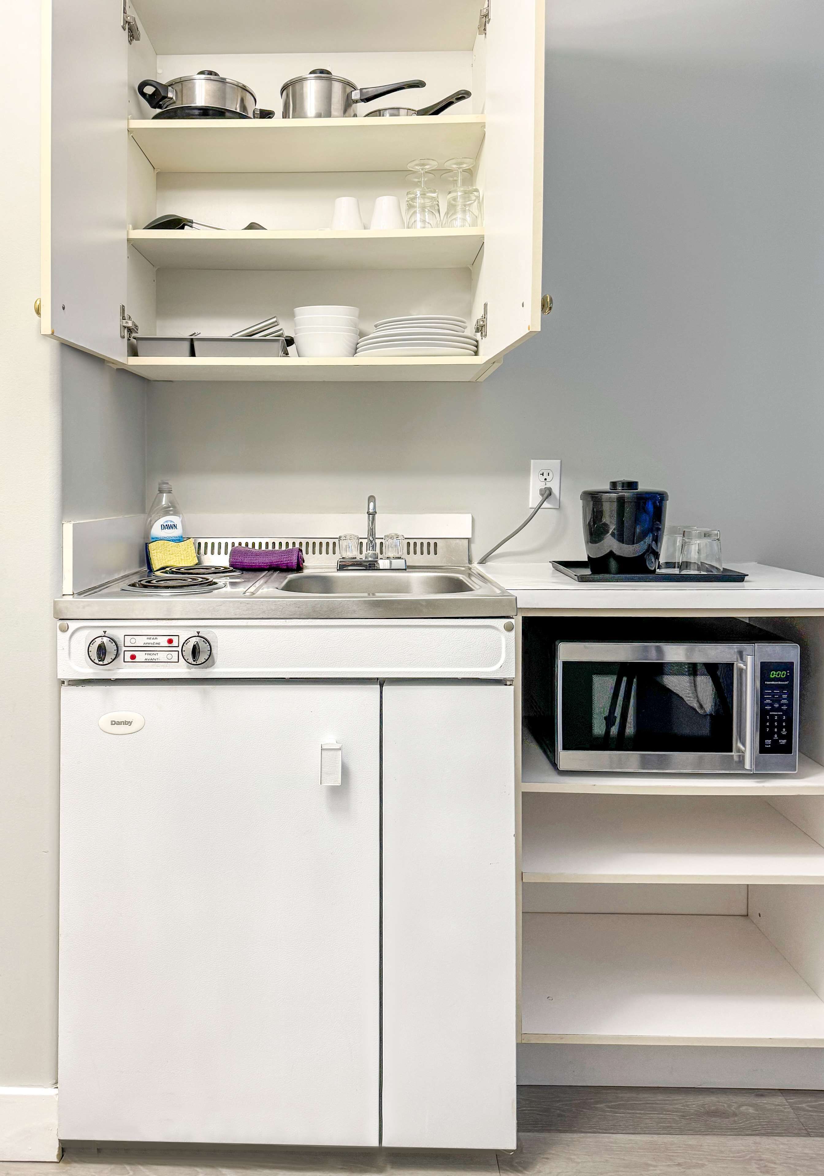 A compact kitchenette with a stove, microwave, and open shelving above containing dishes and cookware.