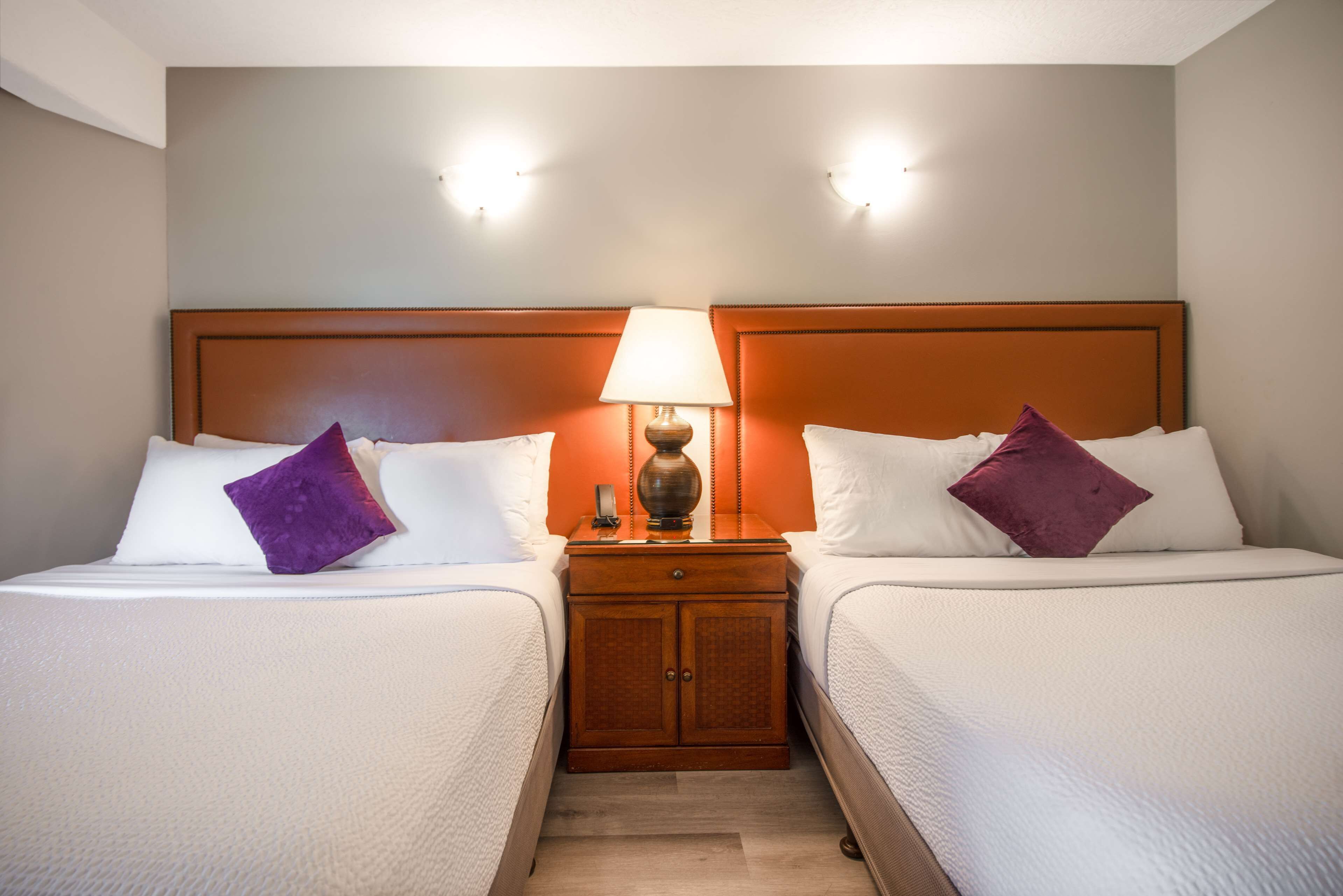 A hotel room with two beds, a nightstand with a lamp, and a wooden headboard.