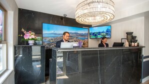 Ein Empfangsschalter mit zwei Mitarbeitern, einem großen Flachbildfernseher, der eine Stadtansicht zeigt, und einer dekorativen Kristallleuchter.